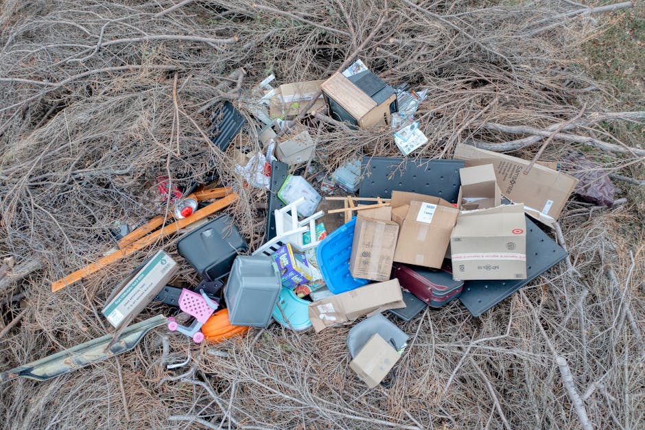 An outdoor scene displaying a pile of miscellaneous rubbish partially surrounded by dry, leafless branches and twigs. The waste includes various cardboard boxes, some flattened and others still assembled, along with plastic containers in different colors such as blue, grey, and pink, and a black plastic tray. There are also small household items like a red soda can, a pink plastic basket, and a white plastic crib or baby chair. A piece of orange timber and a long, thin wooden slat are visible among the debris, with some items leaning on or intermingled with the branches. The background features more dry plant material with no significant structures, indicating the rubbish has been dumped on a natural, unkempt area, possibly a woodland or neglected outdoor space. The overall scene suggests illegal dumping or an abandoned collection of household waste needing professional removal, possibly by services like Waste Clearance Paddington for alternative disposal or on-site clearance solutions.