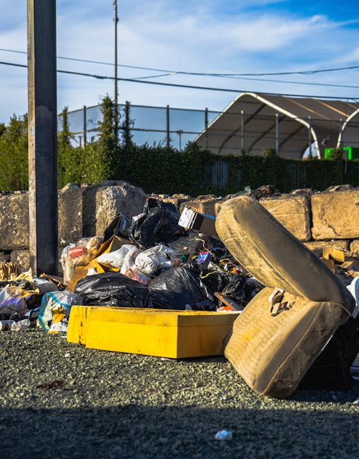 A pile of dumped rubbish situated on a paved surface near a stone wall, consisting of black plastic garbage bags, a large yellow plastic container, an old, dirty car tyre, and broken miscellaneous waste items. In the background, there is a metal fence, some greenery, and a large fabric-covered outdoor structure. The scene is illuminated by natural daylight, with electrical wires overhead and a blue sky visible. The debris appears to be discarded household or garden waste, and the setting suggests an outdoor area where private waste clearance by services like Waste Clearance Paddington might be required to prevent fly-tipping and maintain environmental standards.