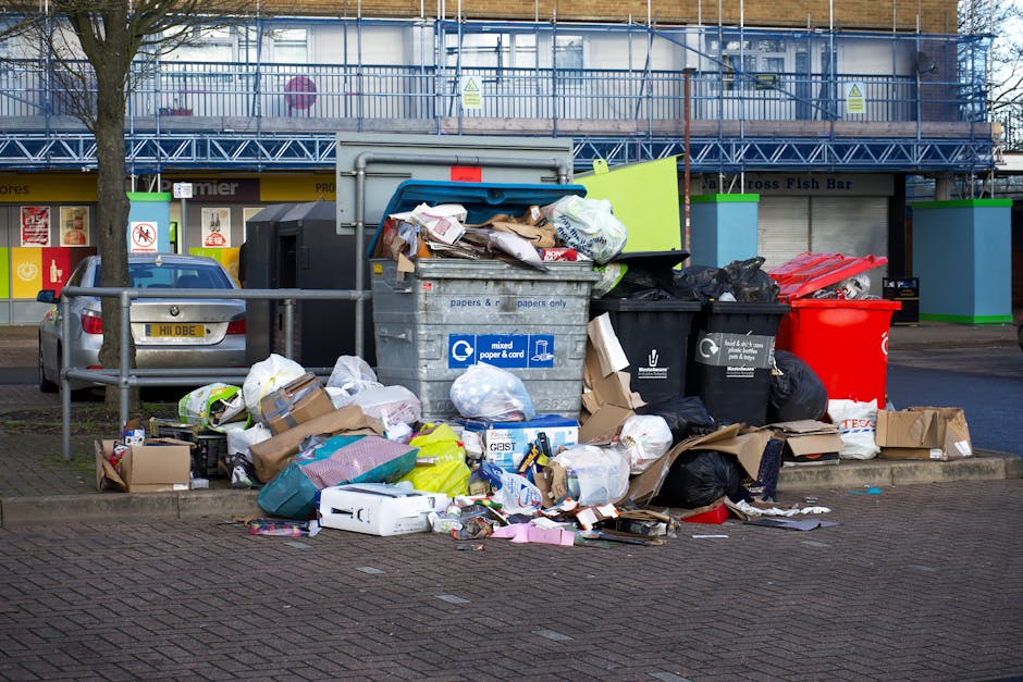 A large collection of mixed waste and rubbish overflowing from several black and red trash bins, along with numerous cardboard boxes and paper bags, are piled on the pavement in front of a commercial building. The metal rubbish containers appear nearly full, with some of the paper and plastic waste spilling onto the surrounding ground. The scene includes a variety of materials such as crumpled paper, cardboard packaging, plastic wrappers, and discarded packaging, with some items leaning against the bins and others scattered across the brick pavement. The background shows a building with retail storefronts, yellow signage, and a blue construction safety barrier or scaffolding, indicating a city or commercial district setting. The natural daylight illuminates the area clearly, and a nearby tree partially frames the scene on the left side. This image depicts typical private waste accumulation awaiting collection, relevant to independent rubbish removal services like those offered by Waste Clearance Paddington in the W2 area.