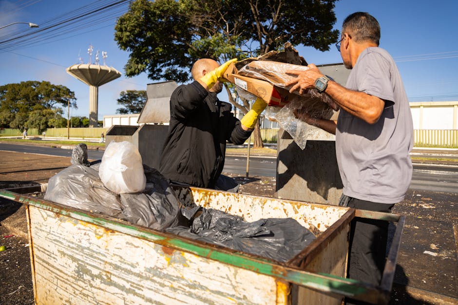 The image depicts two men engaged in rubbish removal or waste collection outdoors on a bright day. One person, dressed in a black jacket and yellow gloves, is seen lifting a cardboard box filled with assorted waste materials, including plastic and paper, from a large, rusted metal container that appears to be a skip or open-top waste bin. The other individual, wearing a light grey T-shirt and wristwatch, is assisting by holding the box steady, ensuring the contents are safely transferred into the container. Surrounding the scene is a paved area with a roadway in the background, featuring parked cars and a row of trees providing green foliage. A distinctive water tower or elevated water tank with a fan-like top structure is visible beyond the trees, along with a section of a modest building with a yellow railing. The environment is clean and well-lit by natural sunlight, emphasizing the practical nature of on-site waste handling that Waste Clearance Paddington might support as an alternative to traditional rubbish collection methods.