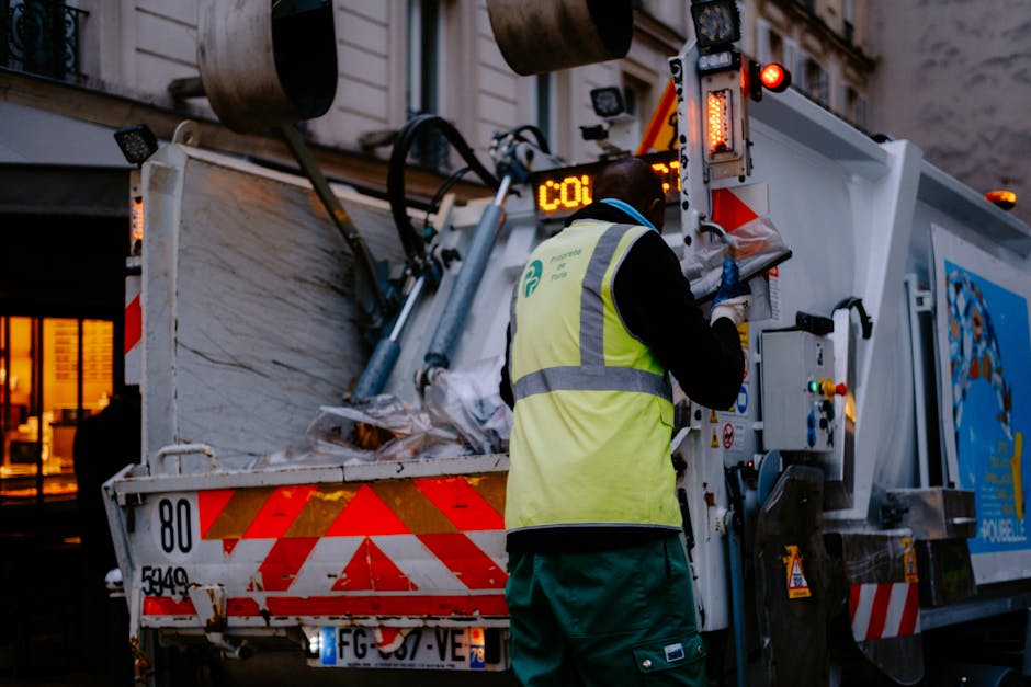 A waste management worker dressed in a high-visibility yellow safety vest with reflective stripes and dark clothing is operating a rubbish collection vehicle on an urban street during dusk or early evening. The worker is standing on the street, facing the back of the refuse truck, which has a white and red chevron pattern on the rear panel. The vehicle's rear section is open, revealing a load of mixed waste materials including paper, plastic bags, and other debris, loosely piled inside. The truck is equipped with hydraulic lifting mechanisms, warning lights, and safety features, with some control panels and buttons partially visible. In the background, buildings with lit windows and a warm ambient glow are visible, indicating a busy city environment. The scene captures the process of private or independent waste collection, relevant to rubbish removal services based in Paddington, with the focus on efficient waste handling and disposal outside residential or commercial premises.