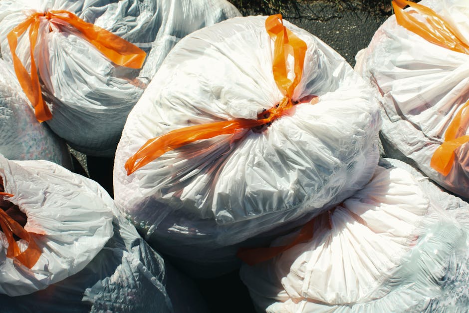 Close-up view of several large white plastic rubbish bags filled with waste, tightly knotted at the top with orange plastic ties. The bags are situated outdoors on a paved surface, with some overlapping each other. The plastic material is semi-transparent, allowing glimpses of mixed household waste inside, and the bags exhibit a slightly crinkled texture under natural daylight. The scene appears to depict a collection of bulky waste or rubbish awaiting collection or disposal, consistent with private waste clearance services like those offered by Waste Clearance Paddington. The environment suggests an outdoor area, possibly a driveway or pavement, used temporarily for waste storage before removal. The overall composition emphasizes the organized gathering of rubbish bags prepared for environmentally responsible disposal or collection, aligning with the concept of alternative waste handling methods outside of standard council services.