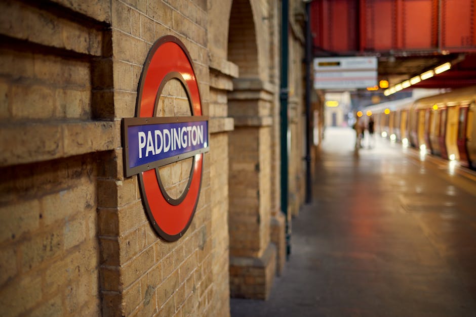 A close-up view of a brick wall at Paddington underground station features a large, circular station sign with a red outer ring, a white inner ring, and a blue rectangular panel displaying the name 'Paddington' in bold white letters. In the background, a platform extends into the distance, with a few blurred figures of passengers walking along it. On the right side, part of an orange and yellow train or subway carriage is visible, with illuminated doors and windows, indicating an active station environment. The platform floor is made of smooth, light-colored surface, and the scene is lit by artificial lighting, creating a warm, welcoming atmosphere typical of busy urban railway stations. This environment may be relevant to discussions about transportation hubs where efficient rubbish removal and waste management are necessary to maintain cleanliness in high-traffic areas, such as those served by private rubbish collection services like Waste Clearance Paddington.