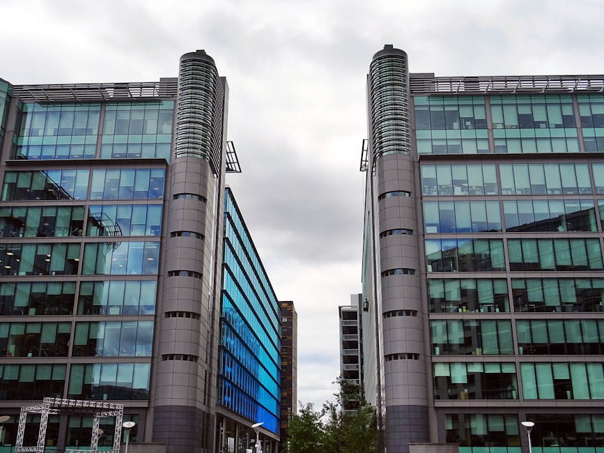 A daytime view of two modern glass-fronted office buildings with curved metallic vertical columns on each side, reflecting the overcast sky and surrounding cityscape in their tinted windows. The buildings are situated on a paved area with some greenery visible in the lower part of the image, and a partial view of additional structures in the background. The glass surfaces display a mix of blue, green, and gray tones, emphasizing their contemporary architectural style. The scene is well-lit despite the cloudy weather, providing a clear and detailed view of the façades, suitable for illustrating topics related to commercial properties or business environments, with an indirect connection to the management and disposal of office waste and rubbish removal services.