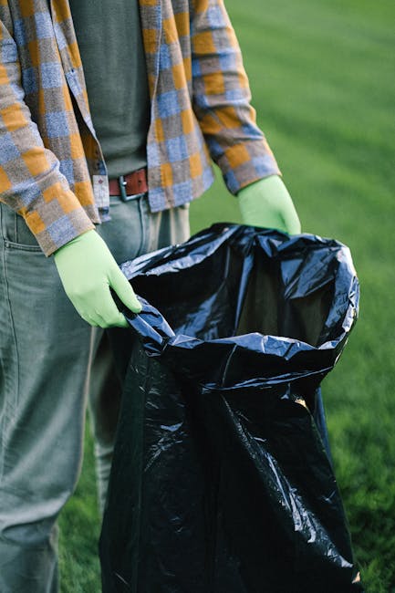 A person wearing a green pair of gloves, a green shirt, and a checkered jacket with shades of yellow, blue, and gray is holding open a large black rubbish bag outdoors on a grassy area. The bag appears to be made of plastic with a shiny texture, and it is empty, ready for waste collection. The background shows a well-maintained lawn with blurred green grass, indicating a park or garden setting. The scene suggests an on-site waste clearance activity, possibly organized by Waste Clearance Paddington, for private rubbish removal or garden waste disposal as part of alternative waste handling options outside of local authority collection. Natural daylight illuminates the scene, emphasizing the textures of the gloves, fabric, and bag material, creating a neutral and professional environment suitable for environmental cleanliness and waste management services.