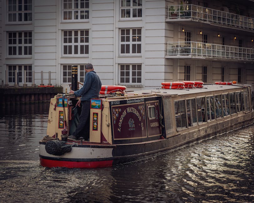 A man standing on the rear deck of a narrow canal boat, which is decorated with colorful panels and has the words 'LONDON WATERBOAT' displayed on its side, is visible in the image. The boat is positioned on a calm river or canal, with slight ripples visible in the water. The boat's roof is equipped with orange life rings and safety equipment, positioned towards the front and center. The man, dressed in dark trousers, a blue jacket, and a flat cap, appears to be operating or preparing the boat. Behind the boat, there is a modern, white multi-story residential building with numerous large windows and balconies, some of which have glass or metal railings. The environment suggests an urban setting with a mixture of waterway transportation and residential infrastructure, indicative of an area where private or independent waste removal or transport services might operate outside of traditional land-based collection methods. The lighting conditions convey either early morning or late afternoon, with soft, diffused natural light illuminating the scene in a neutral, professional manner consistent with an observational depiction of urban waterway activity related to waste management or personnel movements on watercraft.