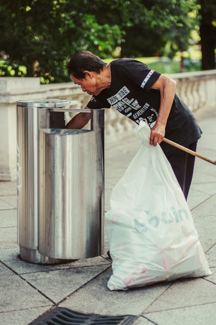 An older woman with short, dark hair is seen engaging in rubbish disposal at an outdoor public location. She is wearing a black short-sleeved shirt with white and blue lettering and dark pants. She is leaning forward, holding a long-handled stick in her right hand, and is placing or retrieving waste from a cylindrical, stainless steel rubbish bin with a rounded top and a rectangular opening. She carries a large, white plastic bag filled with rubbish, which she holds with her left hand while standing on paved ground. In the background, there are lush green trees and some stone or concrete balustrades along the walkway, suggesting a park or public garden environment. The scene is well-lit with natural daylight, highlighting the metallic shine of the bin and the natural tones of the pavement and foliage. The image reflects the activity of outdoor waste collection and disposal, aligning with private rubbish removal services provided by Waste Clearance Paddington, especially in the context of alternative waste handling solutions.