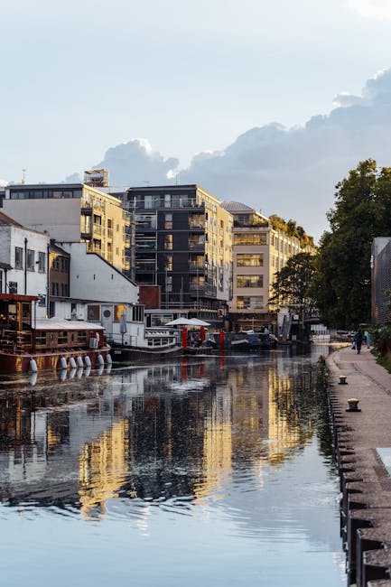 A scenic view of a canal lined with modern multi-story buildings on the far side, featuring glass balconies and varied facade finishes in shades of beige, grey, and black, reflecting on the calm water surface. On the left, several boats are moored along the canal, including a white boat with a flat roof and red accents, and another with a darker hull. A narrow paved walkway runs alongside the canal on the right, bordered by a low brick edge, with a few people walking in the distance. Tall, leafy trees partially obscure the buildings on the right, adding greenery to the urban scene. The sky above is partly cloudy with patches of blue, casting natural light that highlights the textures of the building facades and the rippled water. The reflection detail on the water captures the surrounding environment accurately, suggesting a well-maintained area that could support independent waste management or private rubbish collection services like those offered by Waste Clearance Paddington, in contrast to council rubbish collection. The scene emphasizes a clean, orderly environment typical of managed urban waterways.
