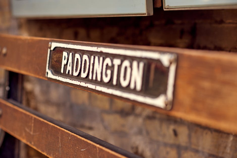 A close-up photograph of a weathered, metallic nameplate displaying the word 'PADDINGTON' in bold, white uppercase letters. The metal surface of the nameplate has visible rust and peeling paint, indicative of age and exposure to the elements. It is mounted on a rustic, wooden slatted structure with a warm brown finish, showing natural grain and slight wear. The background features more wooden elements, possibly part of a fence, gate, or outdoor furniture, with a blurred effect that emphasizes the focus on the nameplate. The overall scene suggests an outdoor setting, perhaps an entrance to a property or an old storage area, relating to independent or private waste handling and rubbish clearance. The environment is naturally lit, with soft shadows and gentle highlights on the metallic and wooden surfaces, aligning with a professional waste management perspective accessible for rubbish removal services.