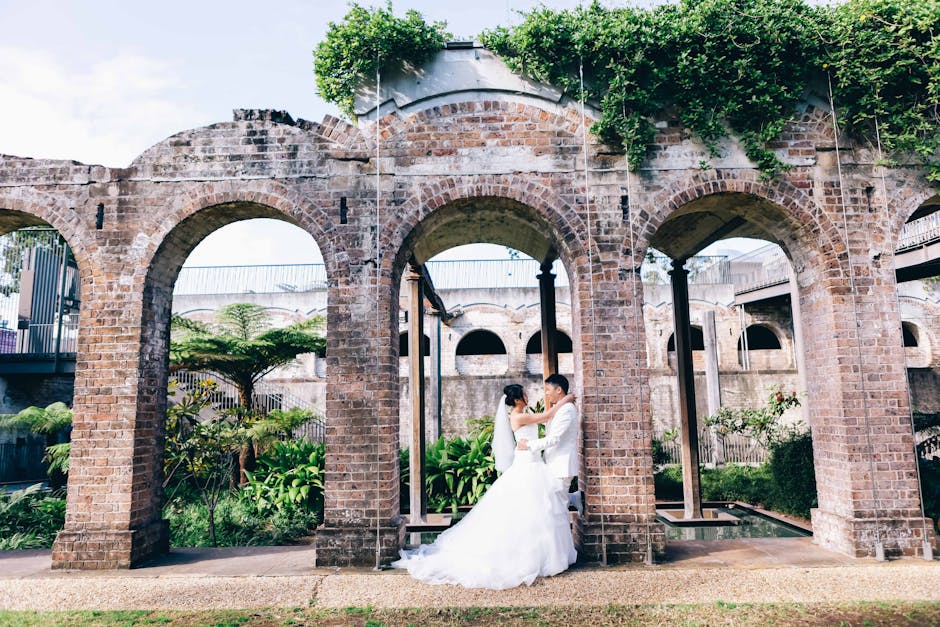 A bride and groom standing close together within a brick archway of a historic outdoor structure, surrounded by lush green foliage and plants. The bride, dressed in a white wedding gown with a veil, faces the groom who is also dressed in white. The scene is set in a garden or courtyard area with additional brick arches in the background, and sunlight filtering through the trees creates a bright, natural atmosphere. The brickwork appears aged with a weathered texture, and the greenery atop the structure adds a natural finishing touch. The setting provides an open-air, rustic environment that can be associated with alternative waste handling or on-site clearance in a natural or outdoor context, subtly aligning with the theme of independent collection and private disposal options. Waste Clearance Paddington occasionally handles such outdoor clearance, ensuring clean and tidy environments around historic or garden settings.