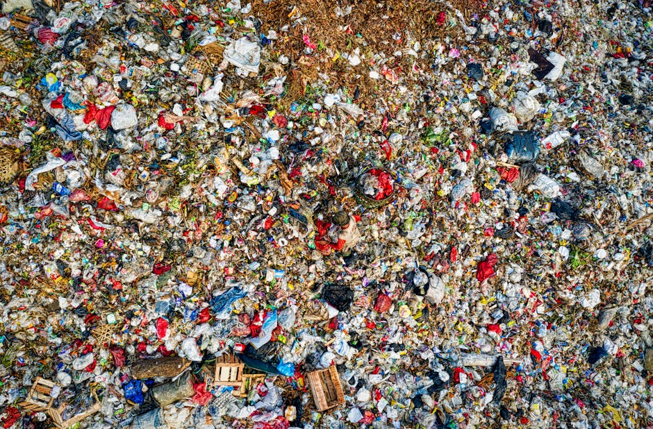 A high-angle view of an extensive and dense landfill site filled with a mixture of waste materials. The image shows layers of tightly packed rubbish, including plastic bottles, paper, cardboard, fabric scraps, and discarded packaging, creating a textured mosaic of various colors, with prominent reds, blues, and browns. The waste appears to be loosely piled, with some items partially buried and others sprawled across the surface. Small wooden pallets are visible amidst the debris, and the overall scene lacks visible machinery or vehicles, suggesting a focus on the waste itself rather than the disposal process. Natural lighting highlights the irregular surfaces and the variety of materials, emphasizing the scale of waste accumulation typical of private rubbish collection and non-recyclable refuse, which may be handled by services such as Waste Clearance Paddington for alternative waste management solutions near Norfolk Square, Paddington.