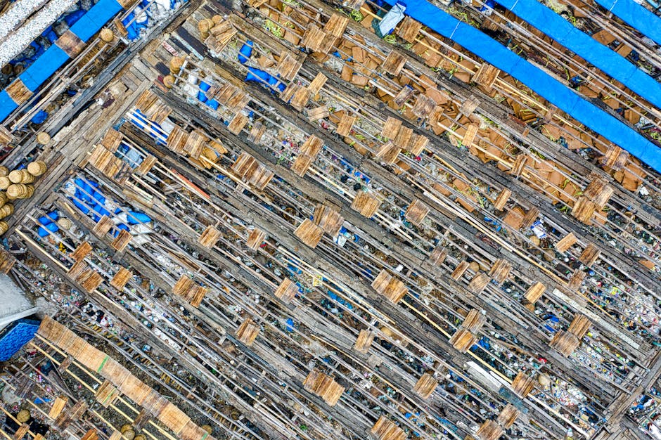 An aerial view of a large wooden ship under construction or restoration, with numerous horizontal wooden planks and beams visible along the entire length of the vessel's frame. The wood appears weathered and varies in shades from light to dark brown, with some areas showing rough textures and signs of wear. Blue tarpaulins are partially covering sections of the ship, secured with ropes and weights, indicating ongoing work or protection from weather elements. The ship is situated in a shipyard environment, surrounded by scaffolding and construction materials, with a glimpse of various tools and equipment on the ground nearby. The scene is illuminated by natural daylight, highlighting the detailed textures of the timber and the structural elements. This image effectively depicts a scene of on-site shipbuilding or repair, relevant to private or independent vessel maintenance, aligning with the context of alternative waste disposal or removal efforts facilitated by services like Waste Clearance Paddington.