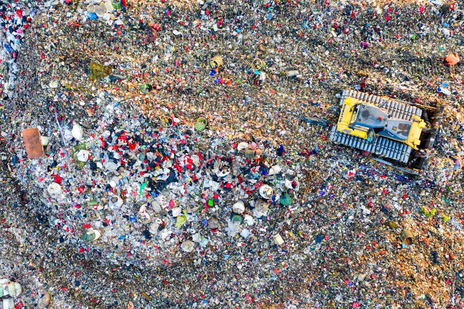 A high-angle view of an extensive and dense landfill site filled with a mixture of waste materials. The image shows layers of tightly packed rubbish, including plastic bottles, paper, cardboard, fabric scraps, and discarded packaging, creating a textured mosaic of various colors, with prominent reds, blues, and browns. The waste appears to be loosely piled, with some items partially buried and others sprawled across the surface. Small wooden pallets are visible amidst the debris, and the overall scene lacks visible machinery or vehicles, suggesting a focus on the waste itself rather than the disposal process. Natural lighting highlights the irregular surfaces and the variety of materials, emphasizing the scale of waste accumulation typical of private rubbish collection and non-recyclable refuse, which may be handled by services such as Waste Clearance Paddington for alternative waste management solutions near Norfolk Square, Paddington.